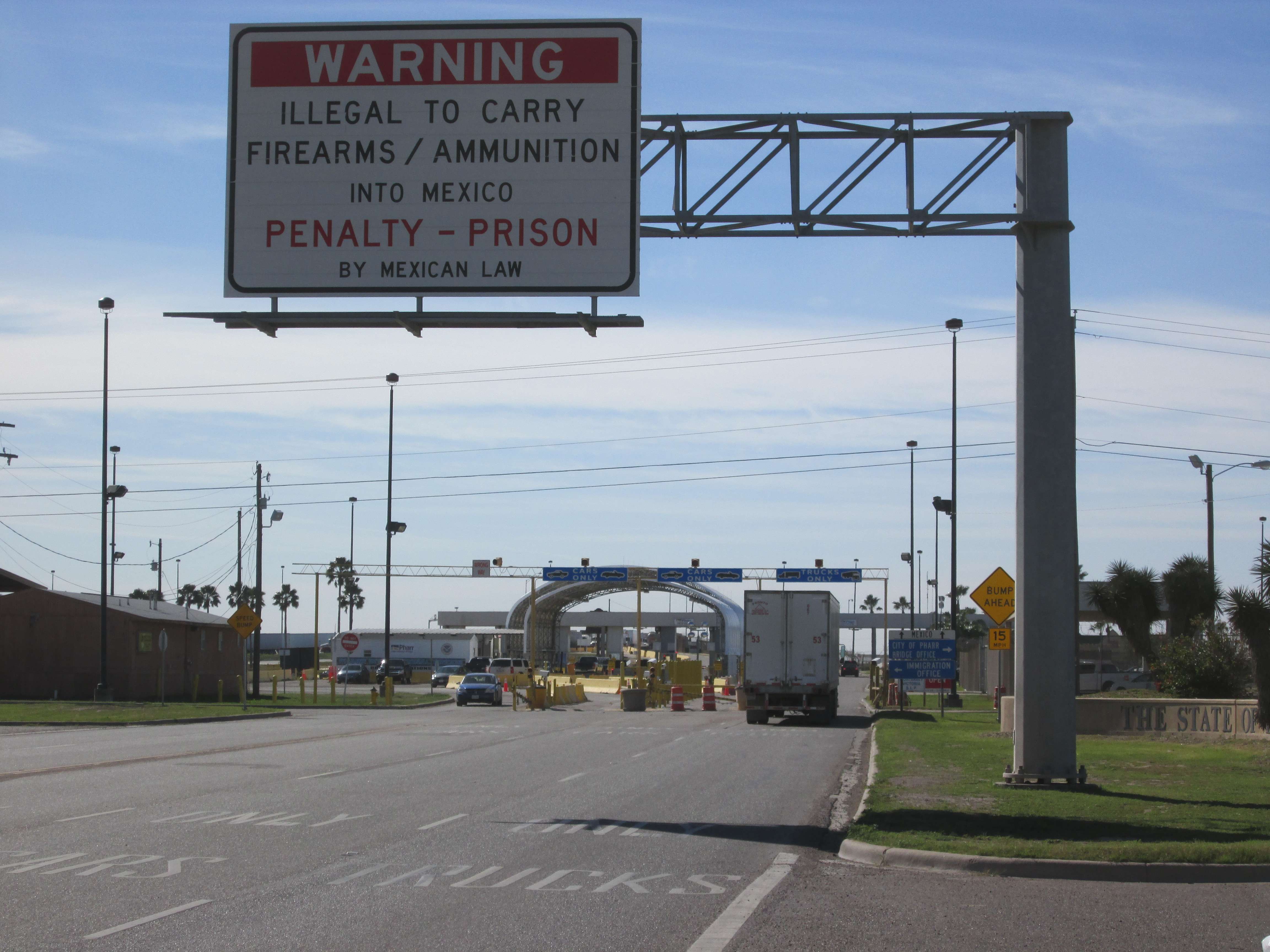 A photo of the Mexican border.  A large sign reads "WARNING | ILLEGAL TO CARRY FIREARMS / AMMUNITION INTO MEXICO | PENALTY - PRISON | BY MEXICAN LAW"